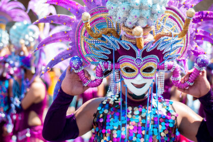 use the keyword , masskara festival in bacolod city and make an alt text for this image. A person adorned in a vibrant, elaborate costume and mask, characteristic of the MassKara Festival in Bacolod City, Philippines, is shown from the chest up. The mask features a smiling face with intricate designs in gold, purple, and blue, framed by large, feathery purple and pink adornments. The costume includes a sequined top in shades of blue, purple, and gold, and the person's arms are covered in purple sleeves, holding the mask in place. The background is blurred, showing other costumed participants, suggesting a festive parade or celebration under bright daylight.