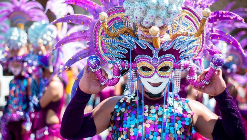 use the keyword , masskara festival in bacolod city and make an alt text for this image. A person adorned in a vibrant, elaborate costume and mask, characteristic of the MassKara Festival in Bacolod City, Philippines, is shown from the chest up. The mask features a smiling face with intricate designs in gold, purple, and blue, framed by large, feathery purple and pink adornments. The costume includes a sequined top in shades of blue, purple, and gold, and the person's arms are covered in purple sleeves, holding the mask in place. The background is blurred, showing other costumed participants, suggesting a festive parade or celebration under bright daylight.