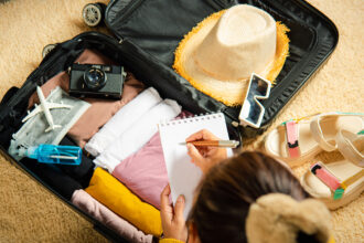 A person, seen from behind, is making a list in a small notebook while sitting next to an open suitcase filled with various travel essentials. Inside the black suitcase, neatly folded clothes in shades of pink, yellow, brown, and white are visible, along with a straw hat, a vintage camera, a small airplane model, a blue spray bottle, and sunglasses. To the right of the suitcase, a pair of light-colored, strapped sandals rests on the beige carpet. This image perfectly illustrates the process of preparing for a trip and figuring out what to pack to the Philippines, ensuring you have all your essentials.
