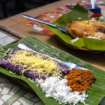 A photo showing Puto Bumbong (purple rice cake) and Bibingka (baked rice cake), served on banana leaves on a mosaic table, representing Filipino Christmas traditions like the post-Simbang Gabi feast.