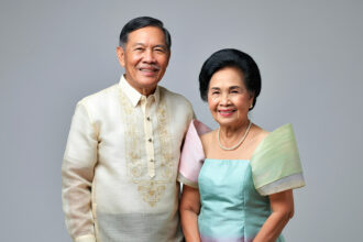 A Filipino man and woman stand side-by-side dressed in elegant Filipino traditional clothing, the man wearing a sheer embroidered Barong Tagalog, and the woman wearing a graceful Maria Clara gown, against a plain backdrop.