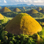 Aerial view of the iconic Chocolate Hills in Bohol, Philippines, showcasing numerous perfectly cone-shaped, brown hills amidst lush green foliage under a partly cloudy sky. This stunning natural wonder is a prime example of the unique and often mysterious fun facts about the Philippines geology and folklore.