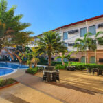 An outdoor resort area with a swimming pool, several palm trees, and dining tables and chairs on a sunny patio, suggesting the amenities of all inclusive resorts Philippines.