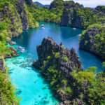 An aerial view reveals the stunning Big Lagoon in El Nido, Palawan, Philippines. Towering, jagged limestone karsts covered in lush green foliage frame a winding waterway that transitions from vibrant turquoise in the shallower foreground, where small boats are anchored, to a deeper indigo blue further back into the lagoon. A few small boats can be seen navigating the calm, clear waters. The scene is breathtaking, highlighting the natural beauty of the region.