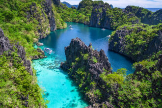 An aerial view reveals the stunning Big Lagoon in El Nido, Palawan, Philippines. Towering, jagged limestone karsts covered in lush green foliage frame a winding waterway that transitions from vibrant turquoise in the shallower foreground, where small boats are anchored, to a deeper indigo blue further back into the lagoon. A few small boats can be seen navigating the calm, clear waters. The scene is breathtaking, highlighting the natural beauty of the region.