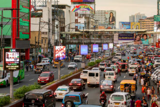 Crowded and busy highway street scene in Metro Manila featuring heavy traffic with cars, jeepneys, buses, and motorbikes, flanked by commercial buildings and large billboards. This image illustrates the dense, bustling nature of the city's main thoroughfares, providing context for travelers researching, "Is Manila safe?" regarding navigating transportation and urban density.