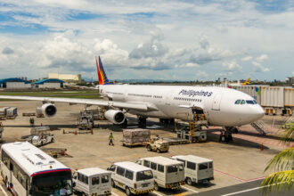 A Philippine Airlines aircraft, identified by "Philippines" on its fuselage and the airline's logo on the tail, is parked at a busy airport gate under a partly cloudy sky. Ground crew and various vehicles, including luggage carts, buses, and vans, are actively working around the plane, suggesting preparations for departure or arrival. The scene makes one wonder about international connections, prompting the question: When will Philippine Airlines resume flights to London?