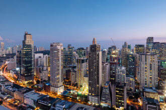 An expansive aerial view captures the vibrant Makati skyline in Manila, Philippines, at dusk. Numerous illuminated skyscrapers of varying heights dominate the frame, their windows glowing with warm light against the deep blue twilight sky. Below, busy streets are marked by streaks of red and white from car lights, indicating the bustling urban environment. The city lights stretch into the distance, showcasing the dense development of this major financial district.
