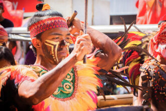 A male performer in face paint and a feathered costume beats a drum with a stick during the Dinagyang Festival celebration.