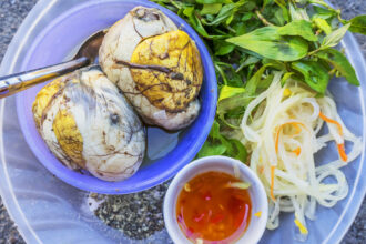 Close-up, overhead view of Balut, the Filipino fertilized duck egg, served in a purple bowl alongside fresh mint leaves, pickled papaya strips, ground pepper, and chili dipping sauce. This controversial delicacy is one of the most talked-about fun facts about the Philippines' unique and adventurous cuisine.