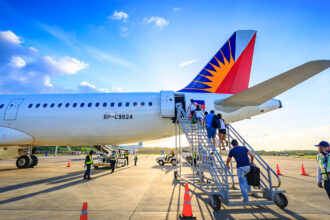 Passengers are boarding a Philippine Airlines aircraft via mobile stairs on a sunny day with a bright blue sky and some clouds. The airline's distinctive sun logo is visible on the tail fin. Ground crew members are also present near the aircraft, and orange cones are set up on the tarmac.