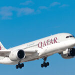 A large Qatar Airways Boeing 787 Dreamliner airplane, painted white with a distinctive purple and maroon tail logo, is captured ascending against a bright blue sky with a few wispy white clouds. As a major international carrier, this aircraft is representative of the types of flights that answer the question: what airlines fly to the Philippines?