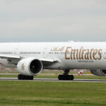 A side-on view of a white Emirates Boeing 777-300ER airplane with the distinctive red, green, and black tail livery, taxiing on a runway. The presence of this major international carrier, which flies long-haul routes, is relevant for those searching for what airlines fly to the Philippines.