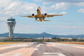 An airplane takes off from a runway, viewed from behind and slightly below, with a tall, modern air traffic control tower standing prominently to the left. In the background, there are low hills and a partly cloudy blue sky, relevant for searching what airlines fly to the philippines.
