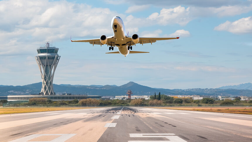 An airplane takes off from a runway, viewed from behind and slightly below, with a tall, modern air traffic control tower standing prominently to the left. In the background, there are low hills and a partly cloudy blue sky, relevant for searching what airlines fly to the philippines.