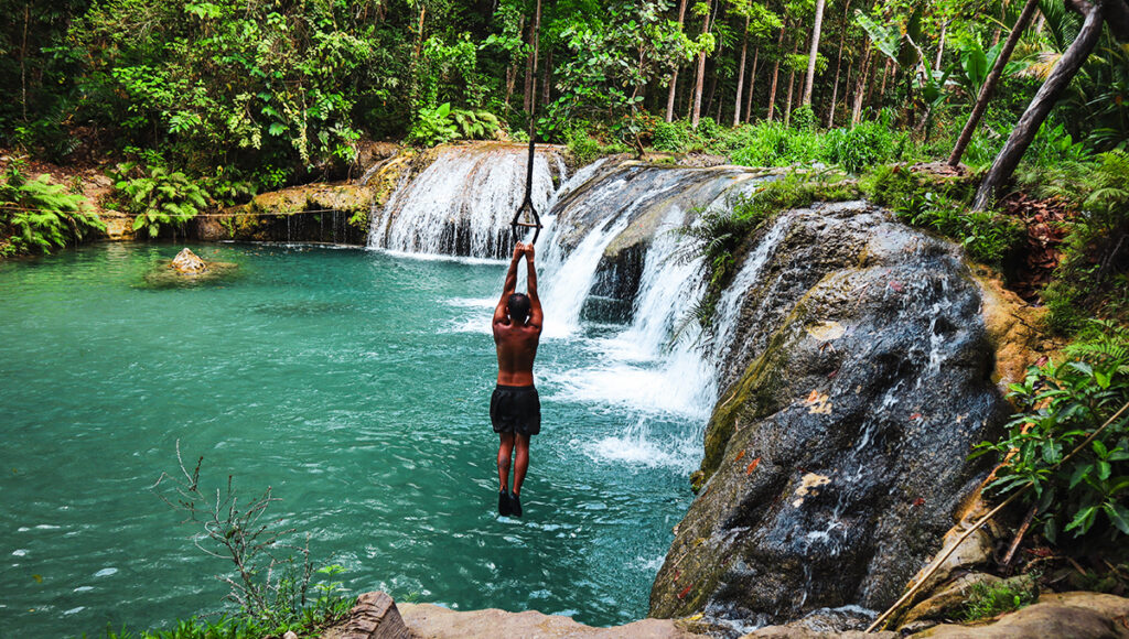 Man swinging on a rope over vibrant waterfalls in the Philippines at Cambugahay Falls with turquoise natural pools in Siquijor.