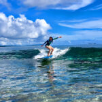 A woman in a long-sleeved swimsuit surfing a clear, turquoise wave at one of the famous Siargao surf spots under a bright blue sky with fluffy white clouds.