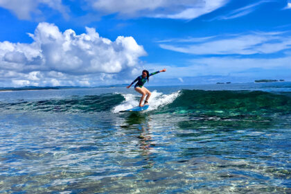 A woman in a long-sleeved swimsuit surfing a clear, turquoise wave at one of the famous Siargao surf spots under a bright blue sky with fluffy white clouds.