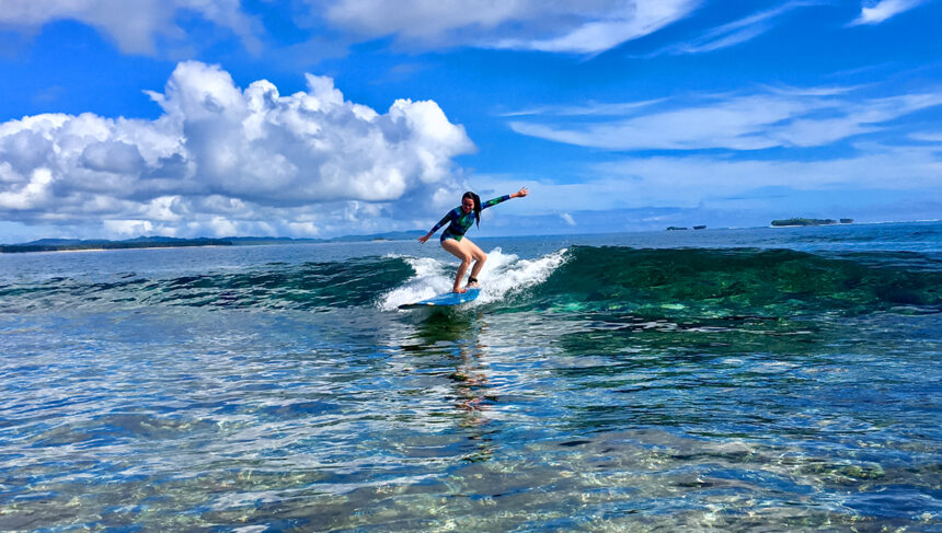 A woman in a long-sleeved swimsuit surfing a clear, turquoise wave at one of the famous Siargao surf spots under a bright blue sky with fluffy white clouds.