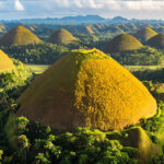 An aerial view of the Chocolate Hills Bohol in the Philippines, showing numerous symmetrical, cone-shaped limestone hills covered in brown and green vegetation under a bright, partly cloudy sky. The foreground features a large, prominent hill with a viewing deck on top, surrounded by lush tropical trees and palm forests.