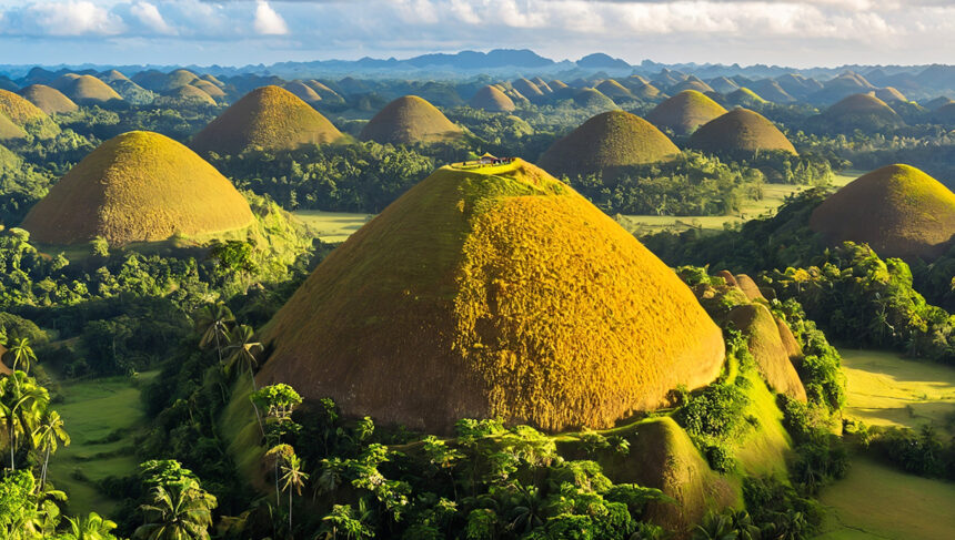 An aerial view of the Chocolate Hills Bohol in the Philippines, showing numerous symmetrical, cone-shaped limestone hills covered in brown and green vegetation under a bright, partly cloudy sky. The foreground features a large, prominent hill with a viewing deck on top, surrounded by lush tropical trees and palm forests.