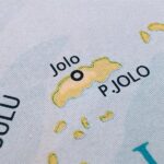 A close-up photograph of a printed map showing Jolo Island, labeled as "P. JOLO," with a black circle indicating the location of the town of Jolo within the Sulu Archipelago.