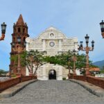 A stone bridge with ornate red lamp posts leading toward the Sanctuario de San Jose, a meticulously reconstructed church and one of the most photographed historical places in Bataan. The church features a white stone facade and a tall, brown brick bell tower, set against a backdrop of mountains and a calm waterway.