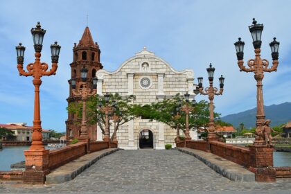 A stone bridge with ornate red lamp posts leading toward the Sanctuario de San Jose, a meticulously reconstructed church and one of the most photographed historical places in Bataan. The church features a white stone facade and a tall, brown brick bell tower, set against a backdrop of mountains and a calm waterway.