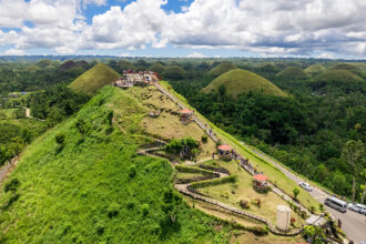 A high-angle view of a main viewing deck atop one of the Chocolate Hills Bohol, featuring a long concrete staircase with several covered rest areas leading up the grassy slope. Tourists are seen on the observation platform and stairs, overlooking a vast landscape of iconic dome-shaped hills and lush green tropical forests under a bright, cloudy sky.