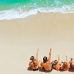 An overhead view of a family of five sitting on a pristine white sand beach and pointing toward the turquoise ocean waves, a common sight at family resorts in Philippines.