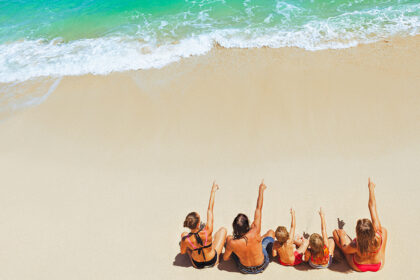 An overhead view of a family of five sitting on a pristine white sand beach and pointing toward the turquoise ocean waves, a common sight at family resorts in Philippines.