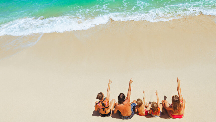 An overhead view of a family of five sitting on a pristine white sand beach and pointing toward the turquoise ocean waves, a common sight at family resorts in Philippines.