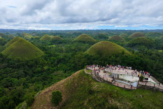 A panoramic view from the summit of one of the Chocolate Hills Bohol, featuring a large, white viewing platform filled with tourists. The platform overlooks a vast valley of symmetrical green hills and dense tropical forests under a dramatic, cloudy sky, capturing the immense scale of the geological formation.