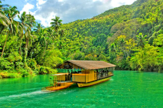A vibrant, eye-level shot of a traditional floating restaurant boat cruising down the bright turquoise waters of the Loboc River in Bohol. The boat is topped with a thatched roof and filled with passengers, while the riverbanks are lined with lush, towering palm trees and dense jungle foliage under a sunny sky.