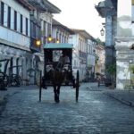 A traditional horse-drawn carriage, known as a kalesa, traveling down the historic cobblestone street of Calle Crisologo. This is one of the most popular things to do in Vigan, showcasing the well-preserved Spanish colonial architecture and vintage street lamps at dusk.