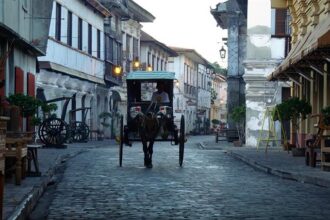 A traditional horse-drawn carriage, known as a kalesa, traveling down the historic cobblestone street of Calle Crisologo. This is one of the most popular things to do in Vigan, showcasing the well-preserved Spanish colonial architecture and vintage street lamps at dusk.