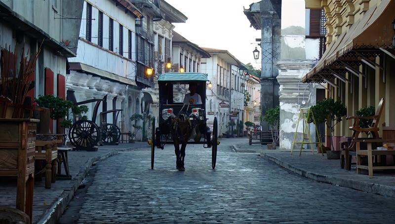 A traditional horse-drawn carriage, known as a kalesa, traveling down the historic cobblestone street of Calle Crisologo. This is one of the most popular things to do in Vigan, showcasing the well-preserved Spanish colonial architecture and vintage street lamps at dusk.