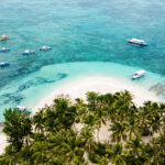 Aerial view of a lush tropical island surrounded by turquoise waters and several white outrigger boats (bangkas) anchored near the shore.