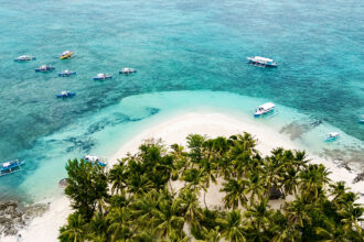 Aerial view of a lush tropical island surrounded by turquoise waters and several white outrigger boats (bangkas) anchored near the shore.