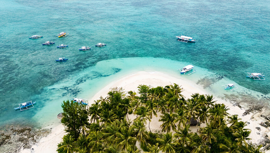 Aerial view of a lush tropical island surrounded by turquoise waters and several white outrigger boats (bangkas) anchored near the shore.