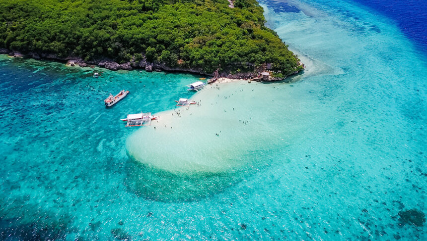 Aerial view of Paliton Beach in Siquijor showing a white sandbar, outrigger boats, and crystal-clear turquoise water under a lush green canopy.