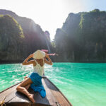 A woman in a traditional conical hat sitting on the bow of a wooden boat, looking out at clear turquoise water and towering karst cliffs under a bright sun.