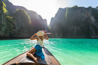 A woman in a traditional conical hat sitting on the bow of a wooden boat, looking out at clear turquoise water and towering karst cliffs under a bright sun.