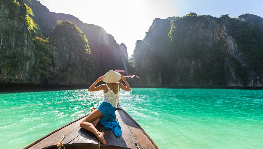 A woman in a traditional conical hat sitting on the bow of a wooden boat, looking out at clear turquoise water and towering karst cliffs under a bright sun.