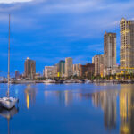 A serene evening view of the Manila Bay skyline reflected in the calm water. Two small sailboats are moored in the foreground, while modern skyscrapers and a yacht club glow under a deep blue twilight sky.
