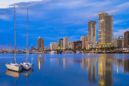 A serene evening view of the Manila Bay skyline reflected in the calm water. Two small sailboats are moored in the foreground, while modern skyscrapers and a yacht club glow under a deep blue twilight sky.