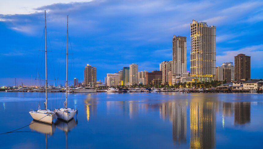 A serene evening view of the Manila Bay skyline reflected in the calm water. Two small sailboats are moored in the foreground, while modern skyscrapers and a yacht club glow under a deep blue twilight sky.
